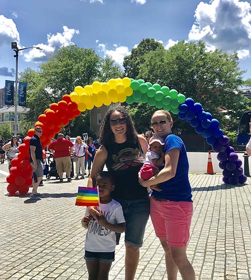 Amie Crawford and family smiling at camera with balloon rainbow in background.