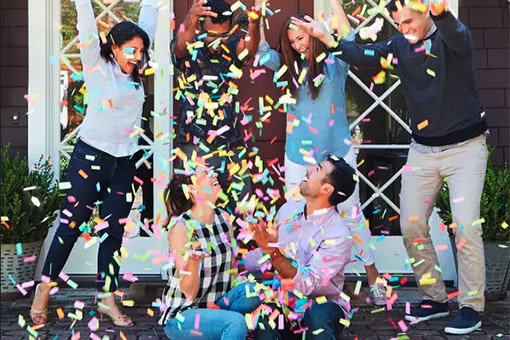 A diverse group of people throwing confetti in the air over a couple sitting on the porch of their new home